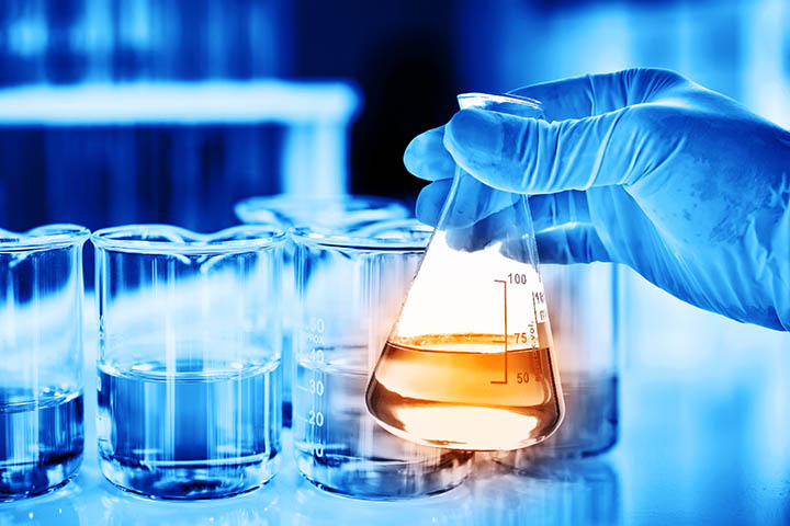 Gloved hand holds an Erlenmeyer flask with orange liquid beside glass beakers in a lab setting.